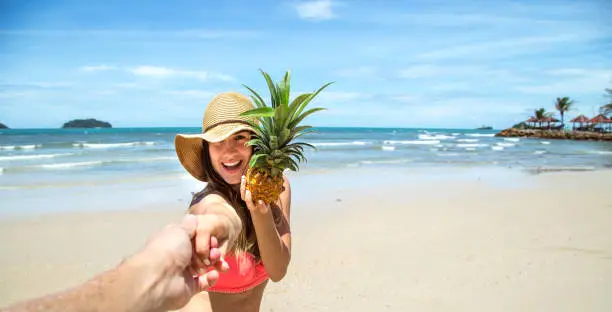 Photo of beautiful girl in swimsuit and pineapple walks on the beach holding the hand of the guy