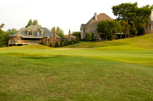 This color image is of Putting Green on Professional Golf Course near Luxury Homes. the putting green is green grass with a sand trap and fairway next to it. the photo is a scenic picture with luxury houses or homes in the background. the picture was taken during the spring or summer and the lighting is natural sunlight taken during the day. 