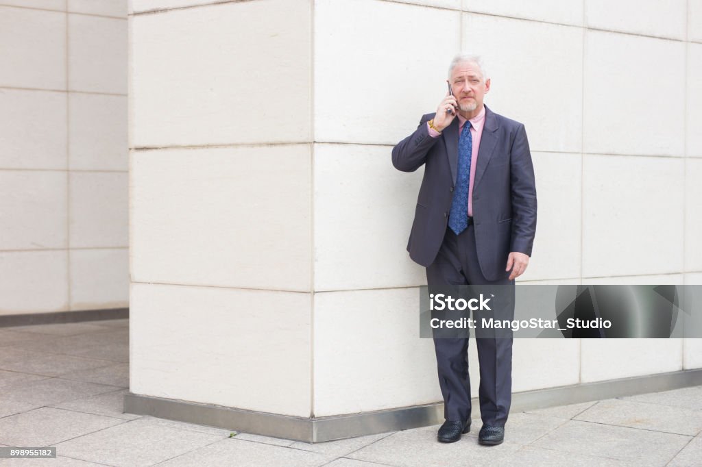 Serious Business Man Talking on Phone Outdoors Portrait of serious senior handsome man looking at camera, talking on smartphone and standing at wall outdoors Adult Stock Photo