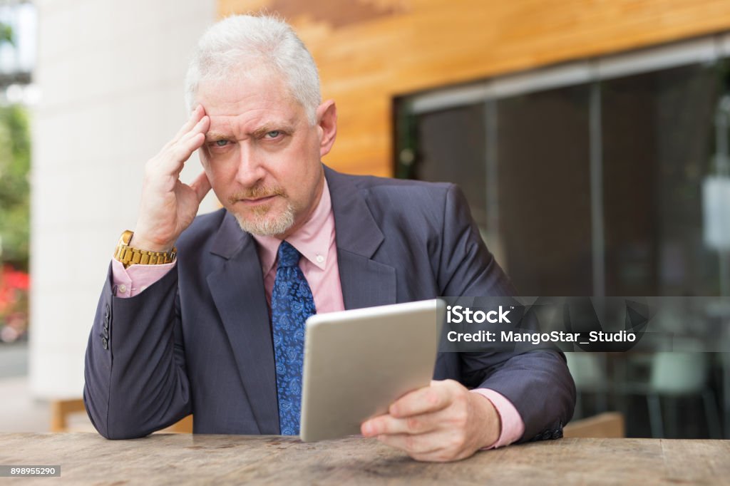 Tensed Business Man with Tablet in Street Cafe Closeup portrait of tensed senior handsome man looking at camera, using tablet computer and sitting at table in outdoor cafe. Front view. Adult Stock Photo