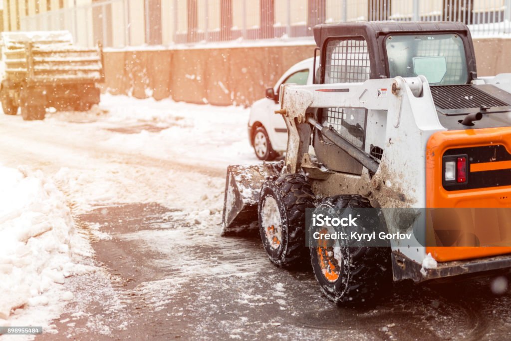 Machine remove snow from a city street Small tractor loader machine clean  and load snow and ice into a truck from a city streets after heavy snowfall Snow Stock Photo