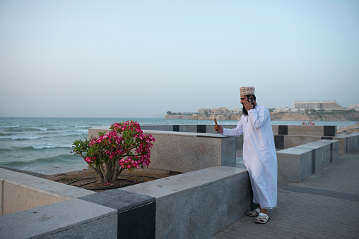 Muscat, Oman - April 20, 2017: A man stops during a walk on the corniche near Al Qurum Beach in Muscat, Oman to take a picture of beautiful blossoms with one mobile phone while talking on another mobile phone.