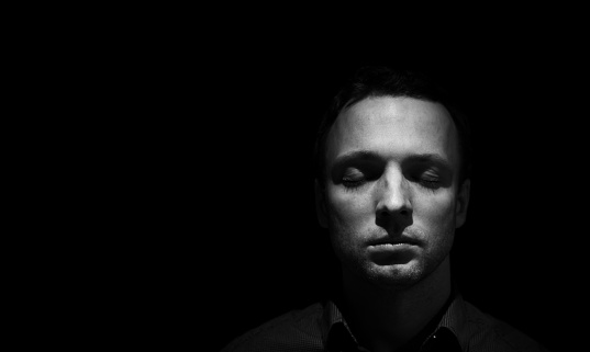 Closeup studio face portrait of young adult European man with closed eyes isolated over black background, low key black and white photo