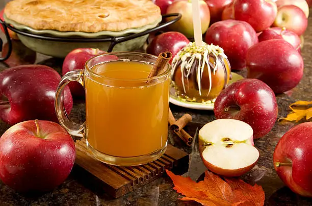 A cup of hot apple cider, caramel apple and apple pie surrounded by fresh fruits