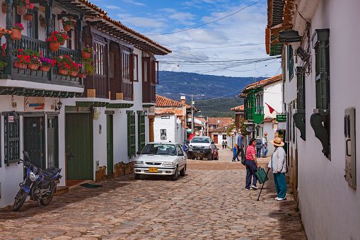 Villa de Leyva, Colombia - November 25, 2017: Looking down hill on one of the cobblestoned streets in the historical 16th Century town of Villa de Leyva in the Department of Boyacá, in the Latin American country of Colombia. The balcony to the left with Geraniums is interesting. The town was founded in 1572 and is located at about 7000 feet above mean sea level on the Andes. In the far background is a section of the Cordillera Oriental of the Andes Mountains. Villa de Leyva was declared a National Monument in 1954 to protect its colonial architecture and heritage. The Town is also famous for being one of the locations for the movie Cobra Verde by Werner Herzog, and the Spanish language Soap Opera, Zorro. Photo shot in the morning sunlight; horizontal format. Copy space. Camera: Canon EOS 5D MII. Lens: Canon EF 24-70mm F2.8L USM.
