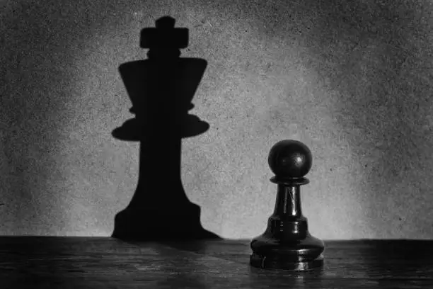 Photo of Chess pawn standing in a spotlight that make a shadow of king with darkness actistic conversion