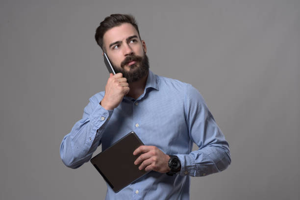 Confident young adult bearded business man talking on the phone while holding tablet computer and looking up at copyspace