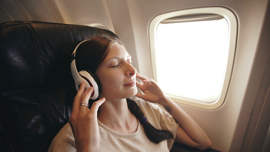 Young woman in wireless headphones listening to music and smiling during fly in an airplane