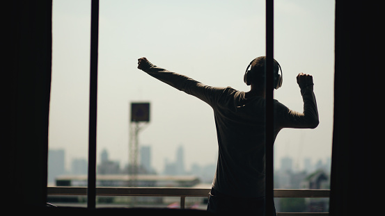 Silhouette of young man dancing ad listening music in wireles headphones stand on hotel balcony