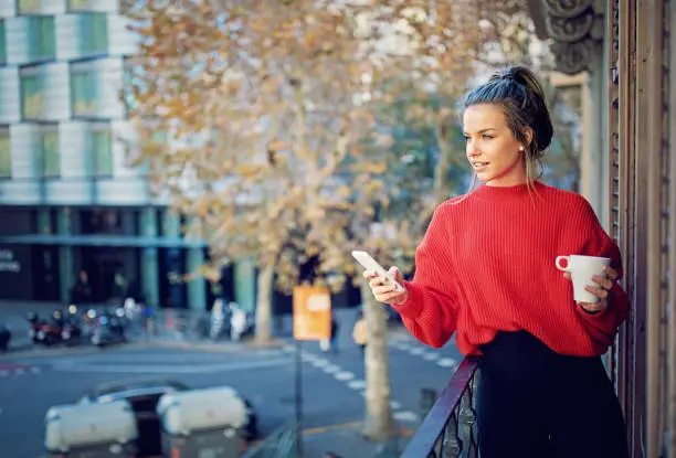 Photo of Young girl is texting and drinking coffee at balcony in the morning