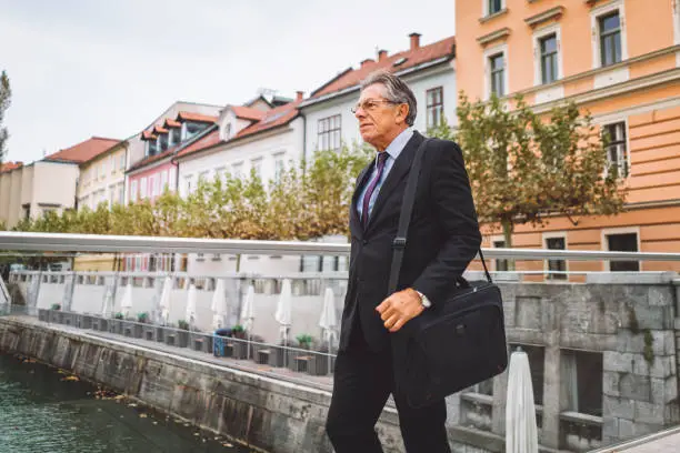 Businessman with specs, happy and successful businessman going to work, walking across the bridge, wearing his elegant business suit, a watch, wearing glasses, staying focused, being serious, making a lot of money, having a lunch break, mature man holding his laptop bag on his shoulder.