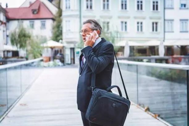 Successful businessman walking around in the city, walking next to the river, holding his work phone, wearing an elegant business suit, glasses, and a watch, wearing a purple tie, talking on the phone, making a lot of money, making new business, mature man has a lot of experiences, confidence is the key to success.