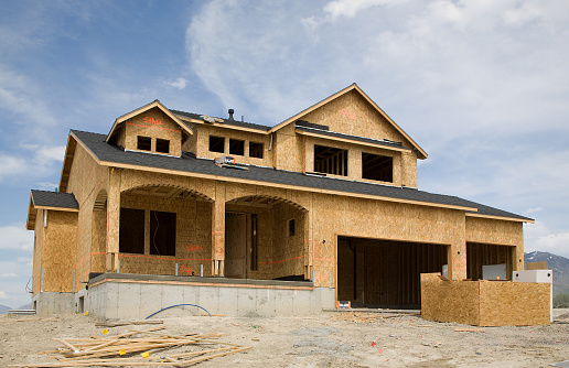 A large luxury home in the process of being built.  The home is framed and has writing on the outside telling what type of finish to complete (stucco, brick, etc.).  On a dirt lot with lots of debris.  Shot against a blue sky.