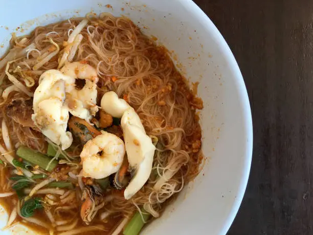 Photo of Thai noodle Tom Yum, spicy seafood noodles soup in a bowl on rustic wooden table, Traditional Thai style food