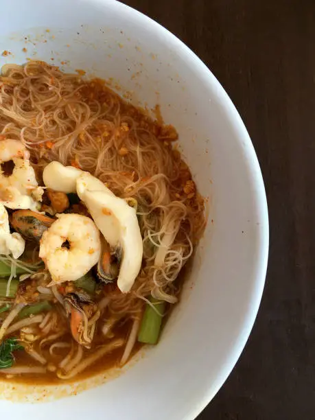 Photo of Thai noodle Tom Yum, spicy seafood noodles soup in a bowl on rustic wooden table, Traditional Thai style food