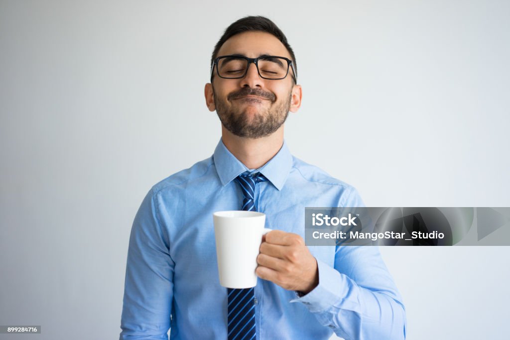 Content Business Man Enjoying Drinking Tea Closeup portrait of content handsome young man holding mug and enjoying drinking tea. Break concept. Isolated front view on grey background. Men Stock Photo