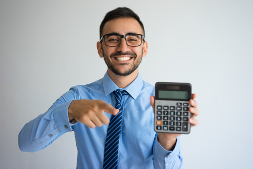 Closeup of smiling handsome young business man holding and showing calculator. Calculation concept. Isolated front view on grey background.