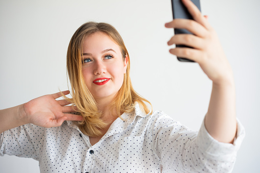Positive attractive businesswoman taking selfie on smartphone against white background. Happy photogenic young woman photographing herself enjoying process. Social media concept