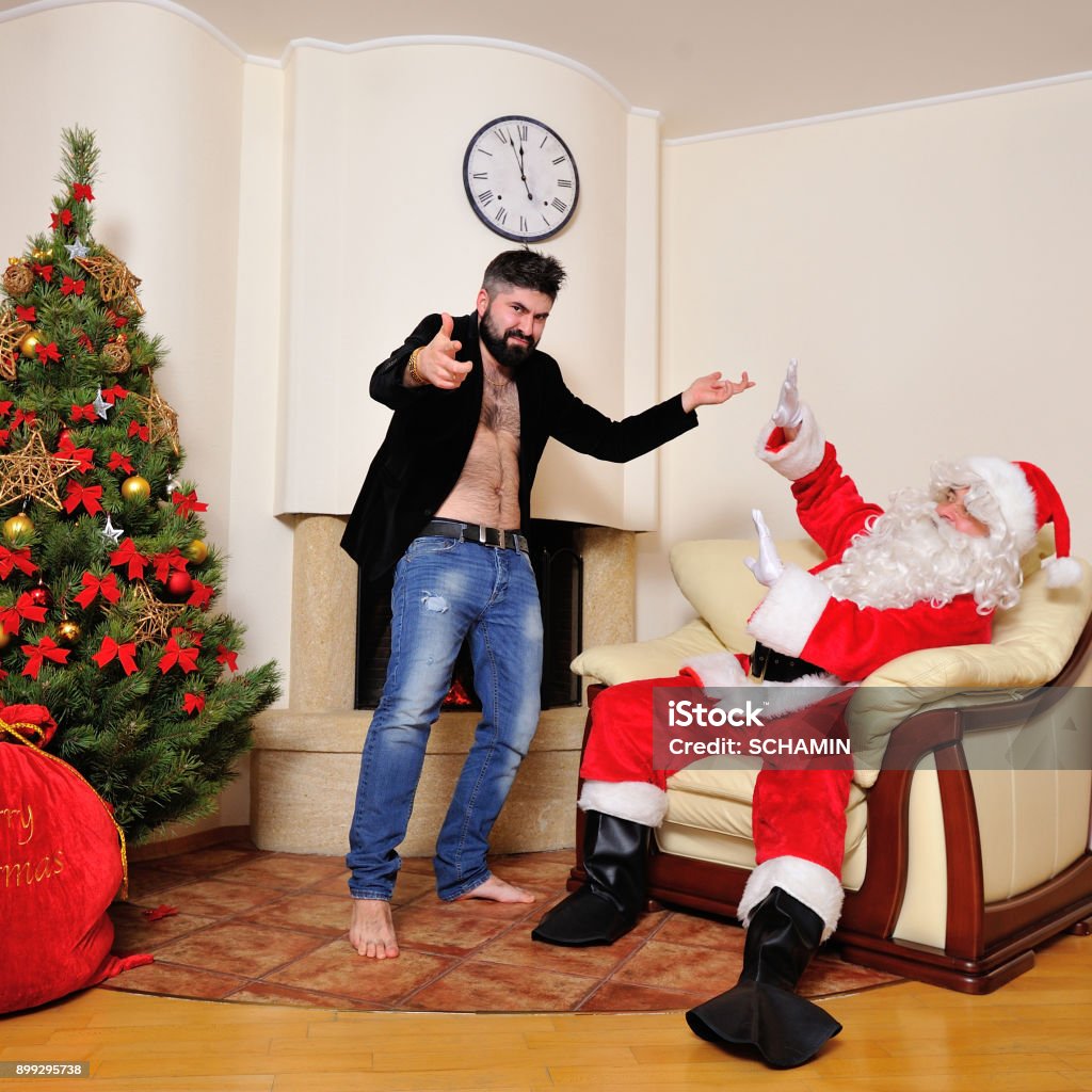 Árbol De Navidad Bolso Del Regalo Chimenea Hombres Vestidos De Papá Noel Y Un Hipster En La Chaqueta De Terciopelo En El Torso Desnudo Baile Juega Al Tonto Mira Loco Foto de Árbol De Navidad Bolso Del Regalo Chimenea Hombres Vestidos De Papá Noel Y Un Hipster En La Chaqueta De Terciopelo En El Torso Desnudo Baile Juega Al Tonto Mira Loco Foto de