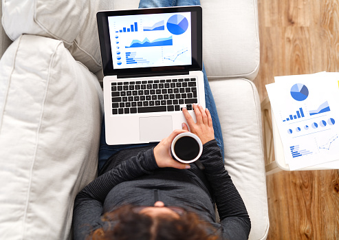 Woman using a laptop and analyzing financial charts while lying on a couch.