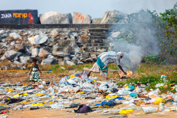 девочка играет в груды мусора в то время как ее мать сжигает его на пляже коллам, штат керала - slum living фотографии стоковые фото и изображения