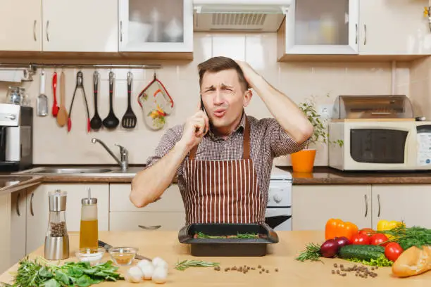 Photo of Perturbed stress young man in apron sitting at table with vegetables, talking on mobile phone, cooking at home preparing meat stake from pork, beef or lamb, in light kitchen with wooden surface.