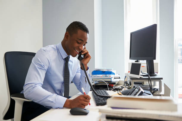giovane uomo d'affari nero che usa il telefono alla scrivania del suo ufficio - giacca e cravatta foto e immagini stock