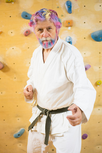 Front view portrait of senior man with rainbow colored hear and beard as rainbow warrior with climbing wall in background.