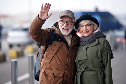 Hello. Waist up portrait of cheerful aged man and woman are standing together outdoors and hugging while looking at camera with joy. Male is rising hand while greeting someone