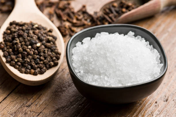 salt and pepper natural salt into black bowl with peppercorn and cloves on wooden table. pink pepper spice ingredient stock pictures, royalty-free photos & images