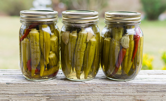 Closeup pickled okra with garlic cloves and red peppers on rustic outdoor table. Useful for grocers, eateries, and homemade foods background.