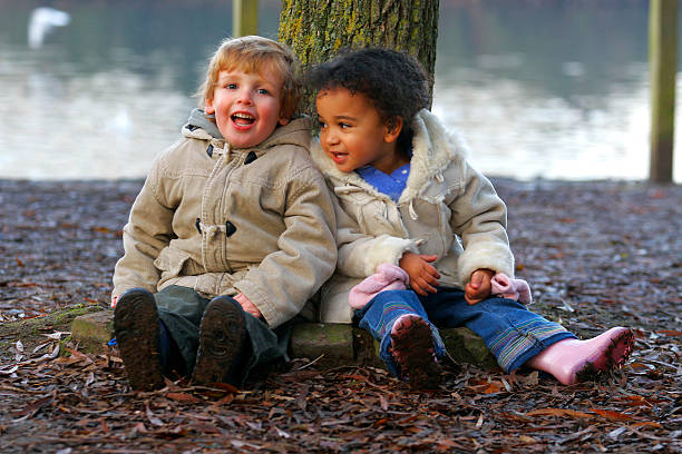 Two Young Children Tired Out Together Leaning On A Tree A little blond boy and a mixed race girl sitting resting at the base of a tree. people covered in mud stock pictures, royalty-free photos & images