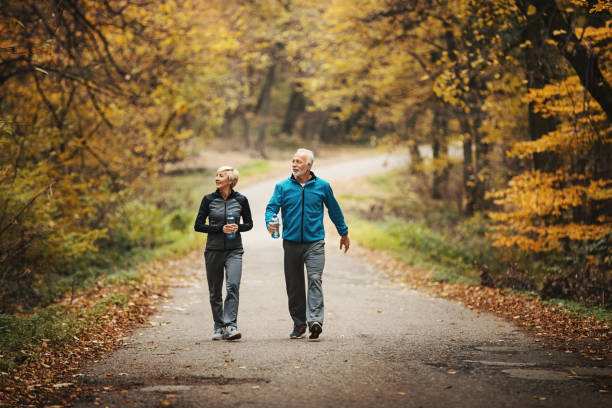 Senior couple power walking in a park. Closeup front view of a senior couple walking in a park after nice jogging workout. They are drinking bottled water and enjoying the view of orange leaves and grass. power walking photos stock pictures, royalty-free photos & images