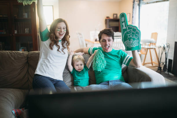 Family Cheering While Watching Football Game A happy family with a young child have fun goofing off while watching a sports game on the television.  They wear their team color and celebrate a win! Touchdown stock pictures, royalty-free photos & images