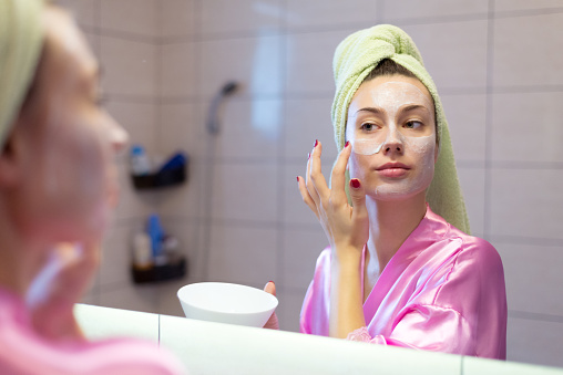 Beautiful young woman putting facial mask on her face in front of the mirror at home.