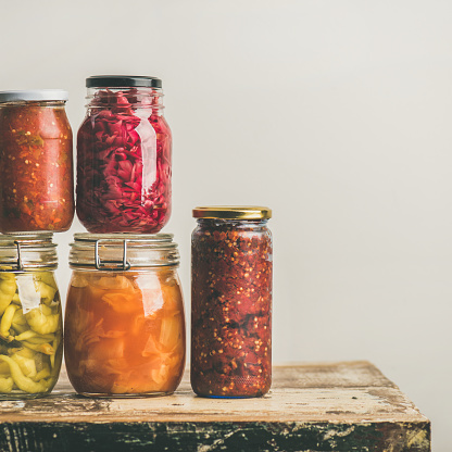 Autumn seasonal pickled or fermented colorful vegetables in glass jars placed in stack over vintage kitchen drawer, white wall background, copy space, square crop. Fall home food preserving or canning