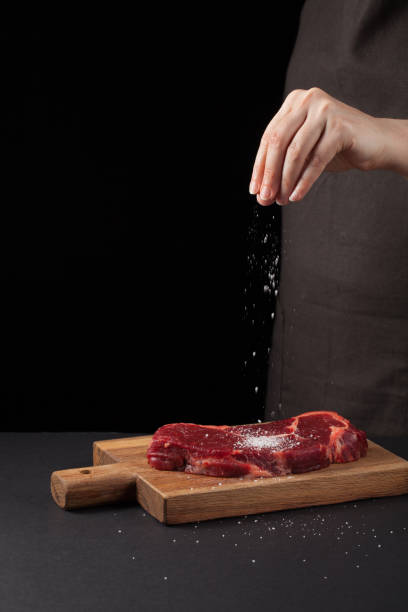 istock Beef steak and salt by chef on the background with free space for text design or logotype menu restaurant. Vertical photo. Food background. Black text area. Copy space 899585702