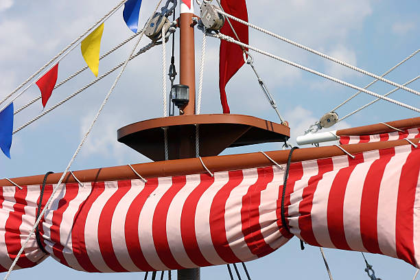 Close-up detail of intricate rigging lines, sails, pulleys and hooks, rope ladders, sail booms and masts as a large sail is being hoisted into place on an old tall ship sailing schooner being readied to cast off and sail at sea.