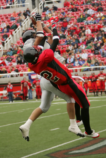 A royalty free DSLR action photo of two football players battling for an airborne ball in a stadium setting. Clad in gray and red uniforms, both players jump and stretch in midair above an astroturf field. The player in gray seems to have the advantage. This image was captured under actual game conditions reflecting competitive intensity. 