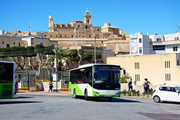Green and white Malta public transport bus in the port area with views towards the citadel and tourists passing by, Victoria (Rabat), Gozo, Malta, Europe.
