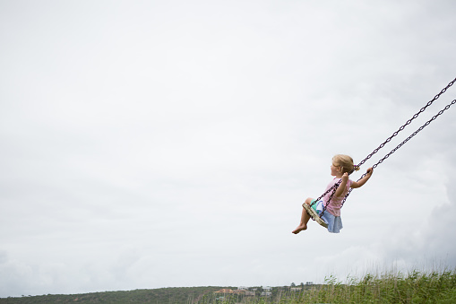 A little girl swings high on a wooden swing on a cloudy day.