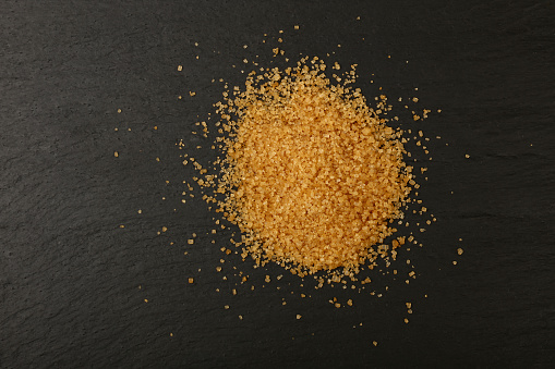 Close up pinch of brown cane sugar spilled on black slate board, elevated top view, directly above