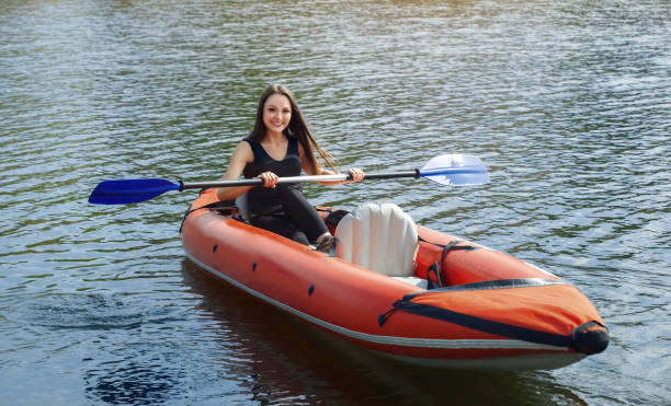 The smiling girl -the sportswoman with longdark hair in blacksportswear rows with an oar on the lake in a red inflatable canoe in a warm summer sunny day The smiling girl -the sportswoman with longdark hair in blacksportswear rows with an oar on the lake in a red inflatable canoe in a warm summer sunny day. Occupation by sports rowing on a kayaking. river swimming women water stock pictures, royalty-free photos & images