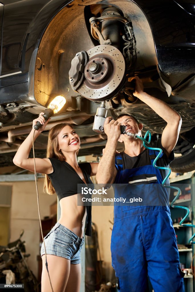 Male car mechanic repairing the car with pneumatic key on hydraulic lift and girl in a frank dressed is standing next to him holding a flashlight. Repairing Stock Photo