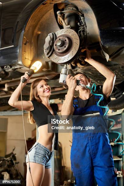 Male Car Mechanic Repairing The Car With Pneumatic Key On Hydraulic Lift And Girl In A Frank Dressed Is Standing Next To Him Holding A Flashlight Stock Photo - Download Image Now