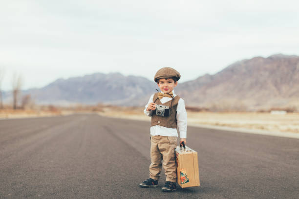 A young boy dressed in an old-fashioned vest, bow tie and driver's cap carries his suitcase on a rural road in Utah, USA. He is ready to go explore the world on his own and take pictures of it while he is gone.