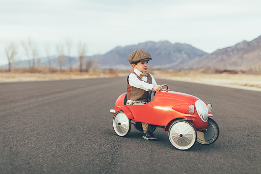 A young boy dressed in vintage vest, bow tie and drivers cap sits in a vintage retro-styled red speedster car on a rural road in Utah, USA. He looks to the right while sitting in the automobile. Plenty of copy space for your concept.
