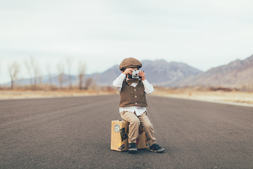 A young boy dressed in an old-fashioned vest, bow tie and driver's cap takes a picture with his camera while sitting on his suitcase on a rural road in Utah, USA. He is ready to go explore the world on his own and take pictures of it while he is gone.