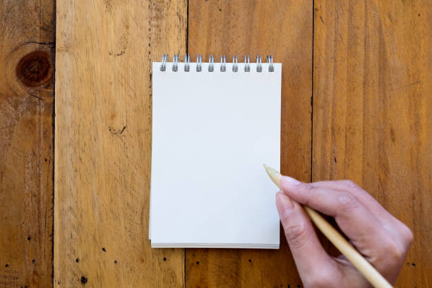 istock Top view image of a hand holding a pen and going to write on a blank white notebook on wooden background 899690668