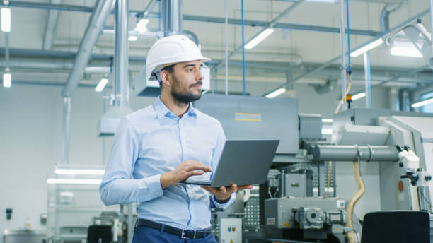 Chief Engineer in the Hard Hat Walks Through Light Modern Factory While Holding Laptop. Successful, Handsome Man in Modern Industrial Environment. Chief Engineer in the Hard Hat Walks Through Light Modern Factory While Holding Laptop. Successful, Handsome Man in Modern Industrial Environment. facilities protection services stock pictures, royalty-free photos & images